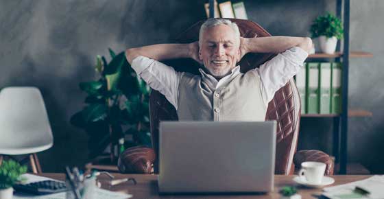 satisfied man in vest sits in front of his laptop computer with hands behind his neck